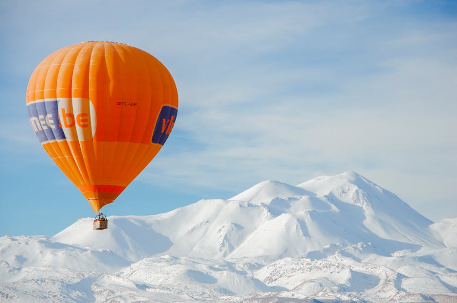 Orange hot air balloon flying over snow-covered mountains under a clear blue sky